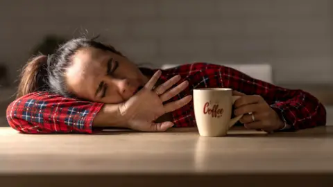 Getty Images A woman in a red checked shirt rests her head on a table next to a cup displaying the word 'coffee'