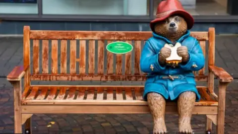 StudioCanal A statue of a Paddington Bear in a red hat and a blue coat holding a sandwich and sitting on a bench.