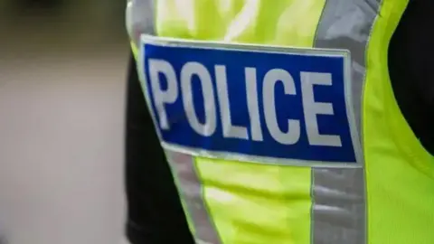 Image shows close-up of a person's back wearing a yellow high-visibility vest with the words "POLICE" written in white and blue across the middle over a black jacket.
