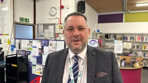 Ollie Conopo/BBC Jason Smithers with short dark hair and beard standing in a library