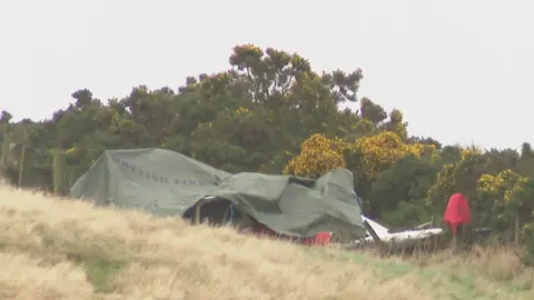 BBC A green tarpaulin has is covering the wreckage. There are trees and bushes behind.