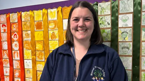 Simon Thake/BBC A woman with dark hair and wearing a navy fleece leans back smiling against a brightly coloured wall covered in children's drawings.