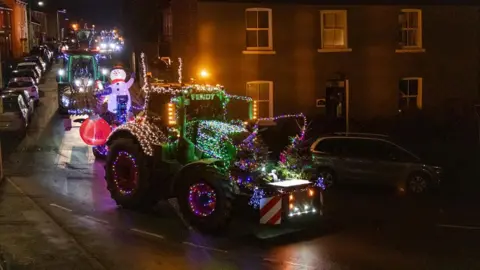 Lina Girling A group of tractors decorated with fairy lights make there way through the streets of a village. The vehicles are travelling in the dark and the surrounding buildings are illuminated by street lights.