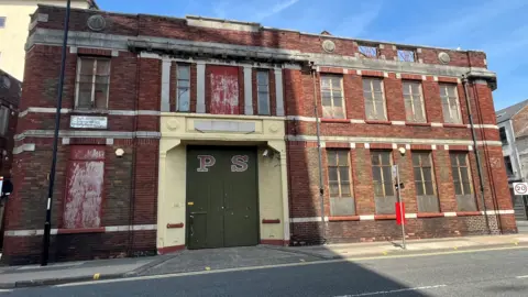 BBC The currently derelict building of the proposed Glassworks site. It is a two-storey, red brick, old factory building with PS written on the green doors.
