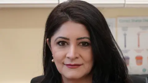 Shabina Asad Qayyum: A woman with dark shoulder-length hair. She is wearing a gold earring in her right ear. Her left ear is hidden by her hair. She is wearing a dark top. A blurred red-and-blue chart is fixed to the wall behind her.
