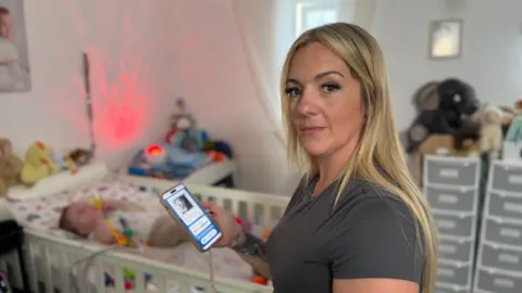 Claire is holding her mobile phone with the app and a picture of Teddy on the screen. She has long blonde hair, is wearing a fitted steel-grey T-shirt and is looking at the camera. In the background, baby Teddy is lying in an ivory-coloured cot, chewing on a teething toy. There are cuddly toys and sensory lights around the head of the cot. 