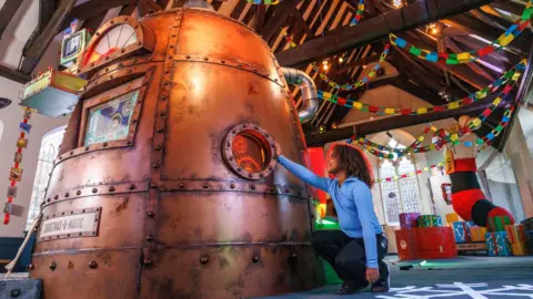 Paul Box A young child wearing dark trousers and a light blue jumper touches the Christmas-o-Matic, a giant copper dome based on Wallace and Gromit's workshop by Aardman Animations. It is inside the Friary at Cabot Circus and behind it festive streamers and toys can be seen