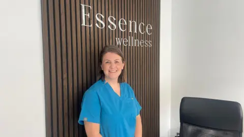 Owner of business in blue scrubs in front of 'Essence Wellness' sign