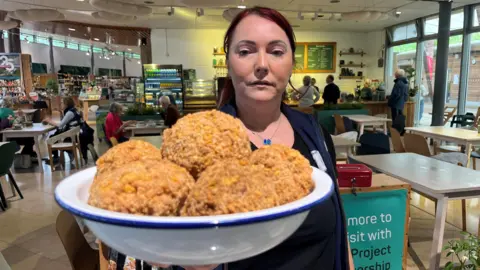 Bernie Williams holding a bowl of large scotch eggs towards the camera. She had tied back red hair and is in a cafe setting. 