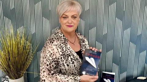 Melanie Graham Mel has short blonde hair, is wearing a leopard-print blouse and has a slight smile on her face. The background wall paper has grey and white stripes. To her left is a vase with yellowish reeds. She is holding a copy of her book Chosen which has a dark cover with Chosen written in red letters and a white paper aeroplane.