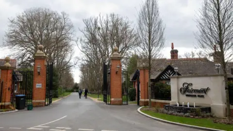 The entrance to the Fairmont Windsor Park Hotel where large pillars in red brick are holding two black metal gates and a straight road continues in to the distance. A sign to the side of the road has the name of the hotel in a script style font.