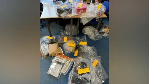 West Mercia Police There is stolen goods in investigation bags. Some are on a blue carpet whilst others are on a table with a brown top and black metal legs. 