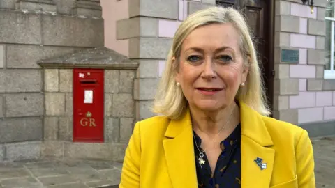 Deputy Helen Miles is wearing a yellow blazer and black top. She has blonde hair and is pictured outside the States Assembly. 