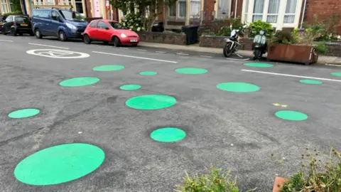 Around a dozen green spots of different sizes across both sides of a residential road, there are planters at either end of the road and parked cars and motorbikes along the opposite side