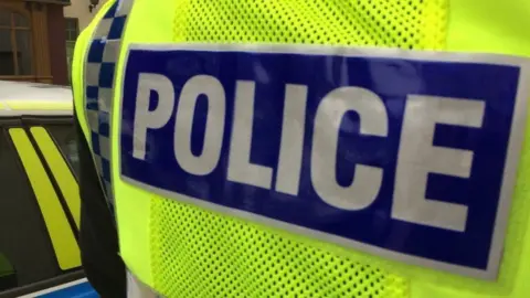 The back of a yellow vest worn over a uniform bears the word 'police'