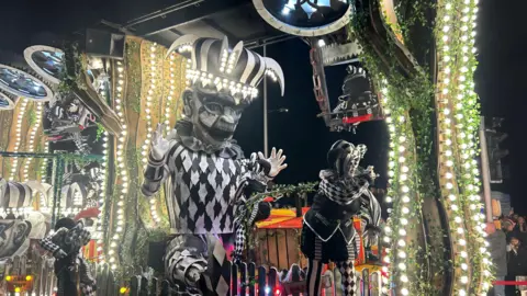 An illuminated float from Weston-super-Mare carnival with large effigies decorated in black and white joker style outfitswith dancers also on the float