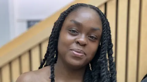 Alex Dunlop/BBC A woman with long black hair in braids smiles at the camera.