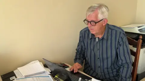 A person wearing glasses and a striped shirt sits at a table using a laptop. Papers, notebooks, a calculator and office equipment are visible around the workspace.