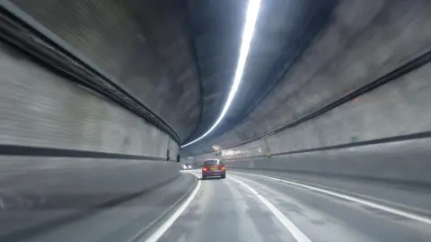 A red car driving through the Rotherhithe tunnel. The tunnel has grey walls and curves to the left. Another car is approaching in the distance, in the opposite direction. 