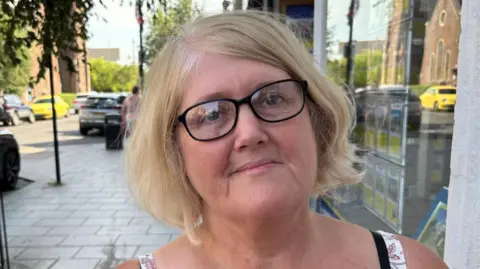 Jodie Halford/BBC Denise Webb has blonde hair and black glasses. She is wearing a vest top on a paved street.