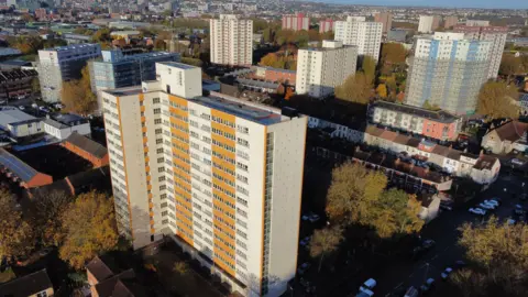 Aerial view of several tower blocks ranging in colour from yellow to blue. Below the buildings are trees and smaller houses.