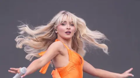 PA Media Sabrina Carpenter faces towards the camera wearing an orange dress. Her hair is spun and she holds the microphone in her hand. 