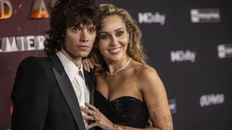 Miley Cyrus, who has long blonde hair and wears a sparkly black strapless dress, smiles on the red carpet as she places a hand - on which she is wearing an engagement ring - on her partner's chest. He has shoulder-length brown curly hair and wears a black suit and whit shirt
