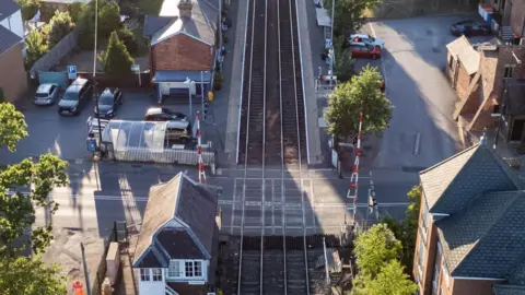 An aerial view of the level crossing that spans the two railway tracks that run down the centre of the frame. Red and white barriers on either side of the crossing are raised. There are cars parked and red-bricked residential houses on both sides of the tracks.