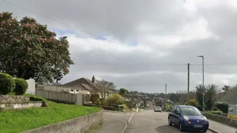 Google A street view of Conway Road in Falmouth. There is a garden with the grass to the left and houses on the right. There are cars parked on the road on the right side and pathways both sides.