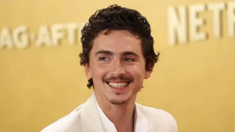 Timothée Chalamet smiling. He is wearing a white shirt and suit jacket. He has dark brown hair. The background behind him is gold.