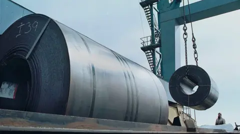 Workers use a crane to load a 60,000 pound coil of steel from Holland onto a railroad car for shipment to Ford Motor Company in Ohio from a warehouse at the South Jersey Port Corporation, March 6, 2002 in Camden, NJ along the Delaware River.