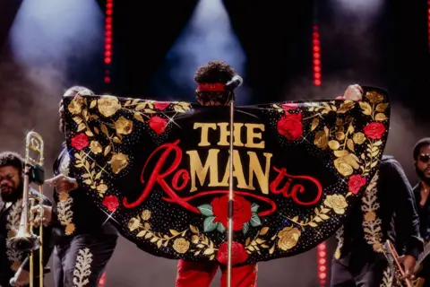 Daniel Ramos Bruno Mars has his back to the audience on stage at Wembley Stadium. He is wearing a black cape that has red and gold roses on it and the words The Romantic. 