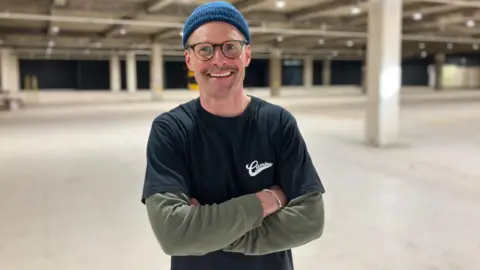 Tim Nokes standing with his arms folded in the empty ground floor of the department store. He is wearing a black short-sleeved T-shirt with a dark green long sleeved T-shirt underneath, black-rimmed glasses and a dark blue beanie hat.