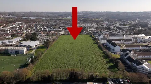 A drone image of the field in St Helier, with a red arrow pointing to more northerly side of the field. There are houses around it.