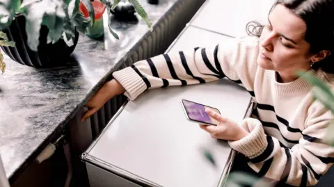 A high angle view of young woman wearing a white and black striped jumper who is checking the temperature of a radiator next to a window. She is looking at a heating app on her smart phone.