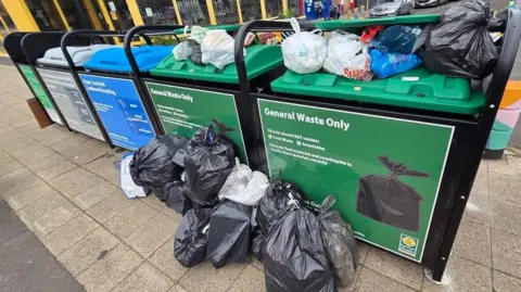 An example of the new bin hubs, consisting of two general waste bins, two recycling bins and a food wastage bin are on the street. The two waste bins are overflowing with rubbish, with a pile of full bin bags also on the ground in front. 