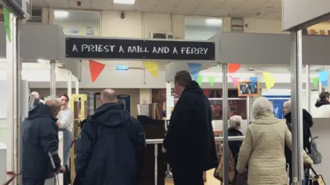 Lara King / BBC People queuing at the A Priest a Mill and a Ferry cafe. There are five people in the queue, each with their backs to the camera. There is multi-coloured bunting up in the cafe. 