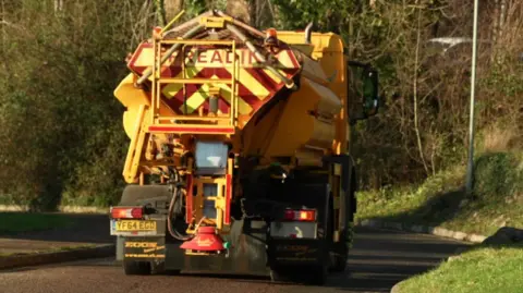 A yellow gritting lorry heads out onto the roads in Devon to spread salt. It is a sunny day and there are grass verges and trees on either side of the road.