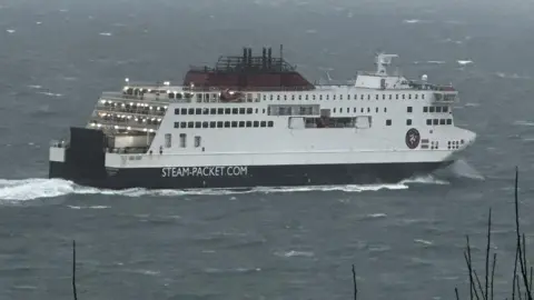 The Manxman ferry sailing out of Douglas Bay. it is a large white ferry with a red funnel and a a black hull that has Steam-Packet.com on the side. The sea is grey and choppy.