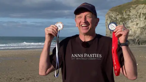 BBC Danny Teare smiling broadly while holding up two bodysurfing medals in either hand.  Danny is wearing a black t-shirt and a navy cap and is standing on a beach with the sea and a cliff in the background.