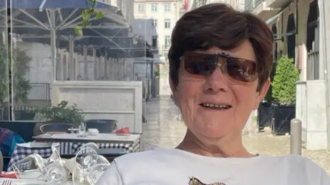 Margaret Crichtonis photographed sitting outside a restaurant on a sunny day. She has short brown hair and wears sunglasses and a white t-shirt.