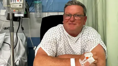 A man with grey hair and glasses and wearing a hospital gown, sits on a chair next to a hospital bed in a ward. Equipment can be seen next to him and a green curtain.