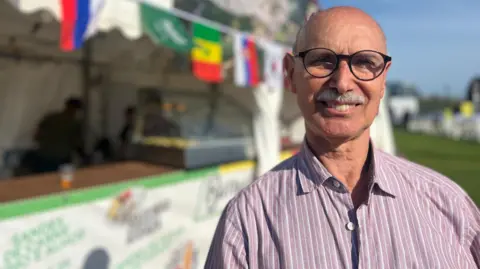 Joao smiles at the camera and behind him there's a food stall with flags over it. He is wearing a pink shirt with white lines on it and has dark rimmed glasses. Joao is bald and has a grey moustache.