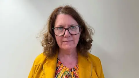 Dr Gilheany is looking at the camera and ia wearing large black framed glasses. She has shoulder length ginger hair and is wearing a yellow jacked and a patterned top.