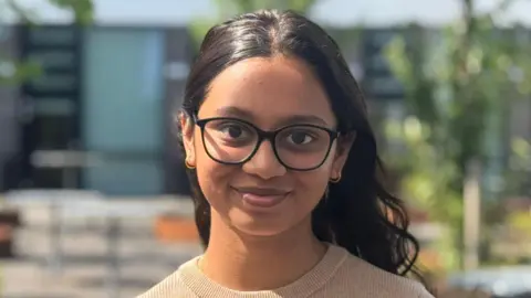 Arya is smiling at the camera. She is wearing a camel-coloured ribbed top and black-framed glasses. She has dark brown wavy hair which is tucked behind her ears and back and she has a small hoop earring in each ear. There are trees and college buildings in the background.