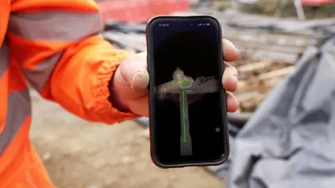 Wessex Archaeology A man holding a mobile phone with the image of a drone down a well on it. 