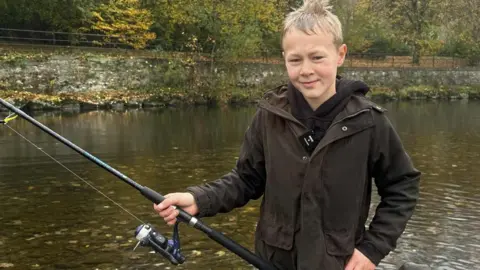 Bailey, a young boy with blonde hair, wears a brown wax coat as he casts a fishing line from his rod into the River Kent. It is a grey rainy day and the visibility is poor.