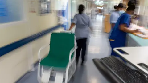 PA Media A blurred image of staff working in a hospital corridor wearing blue scrubs. One staff member is pulling a green wheelchair.