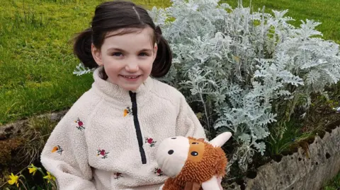 Family handout A little girl with brown hair in pigtails looks towards the camera, smiling. She is wearing a cream fleece with flowers on it and holding a soft toy. She's standing in a garden