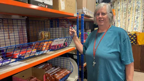 A woman with grey hair is wearing a blue t-shirt. She is stood in a foodbank next to metal shelves which have groceries on them